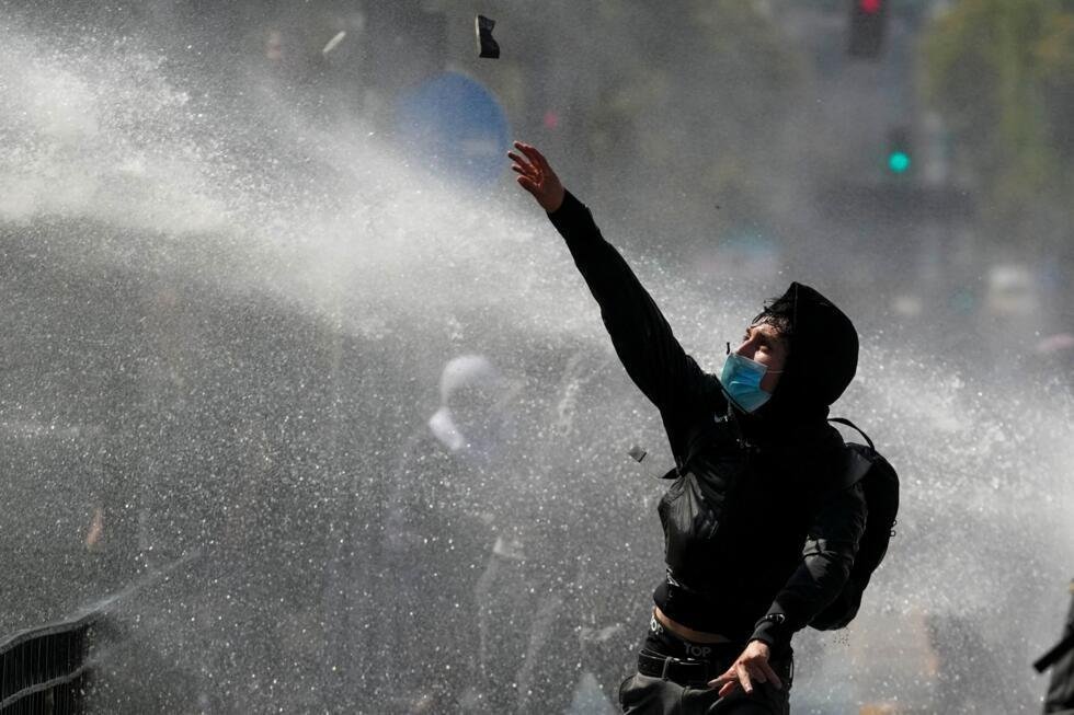 Un joven lanza piedras a la policía, que dispara un cañón de agua en una protesta estudiantil contra la propuesta del gobierno de reducir el presupuesto educativo y contra el aumento de los precios de la gasolina en Santiago de Chile, jueves 26 de marzo de 2026. (