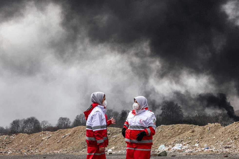 Des femmes membres de la Société du Croissant-Rouge iranien se tiennent près des panaches de fumée d'un incendie dans une raffinerie de pétrole, au nord-ouest de Téhéran, le 8 mars 2026
