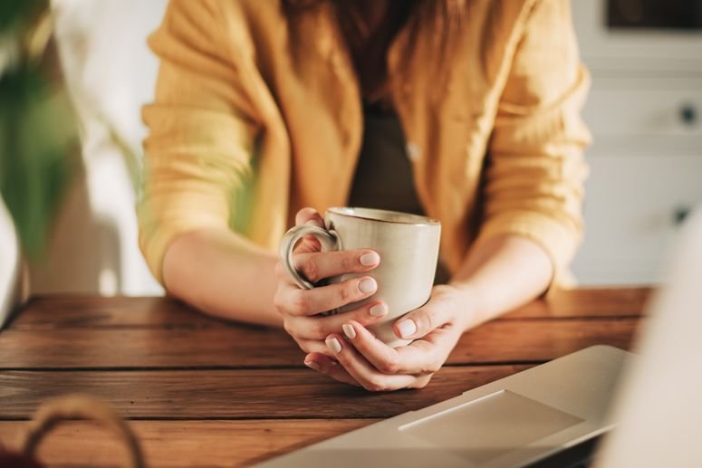 Por la mañana, café para el desayuno y manos femeninas vertiendo agua en una taza den la cocina de casa. Mujer en pijama preparando té o alguna bebida en la encimera para empezar el día.